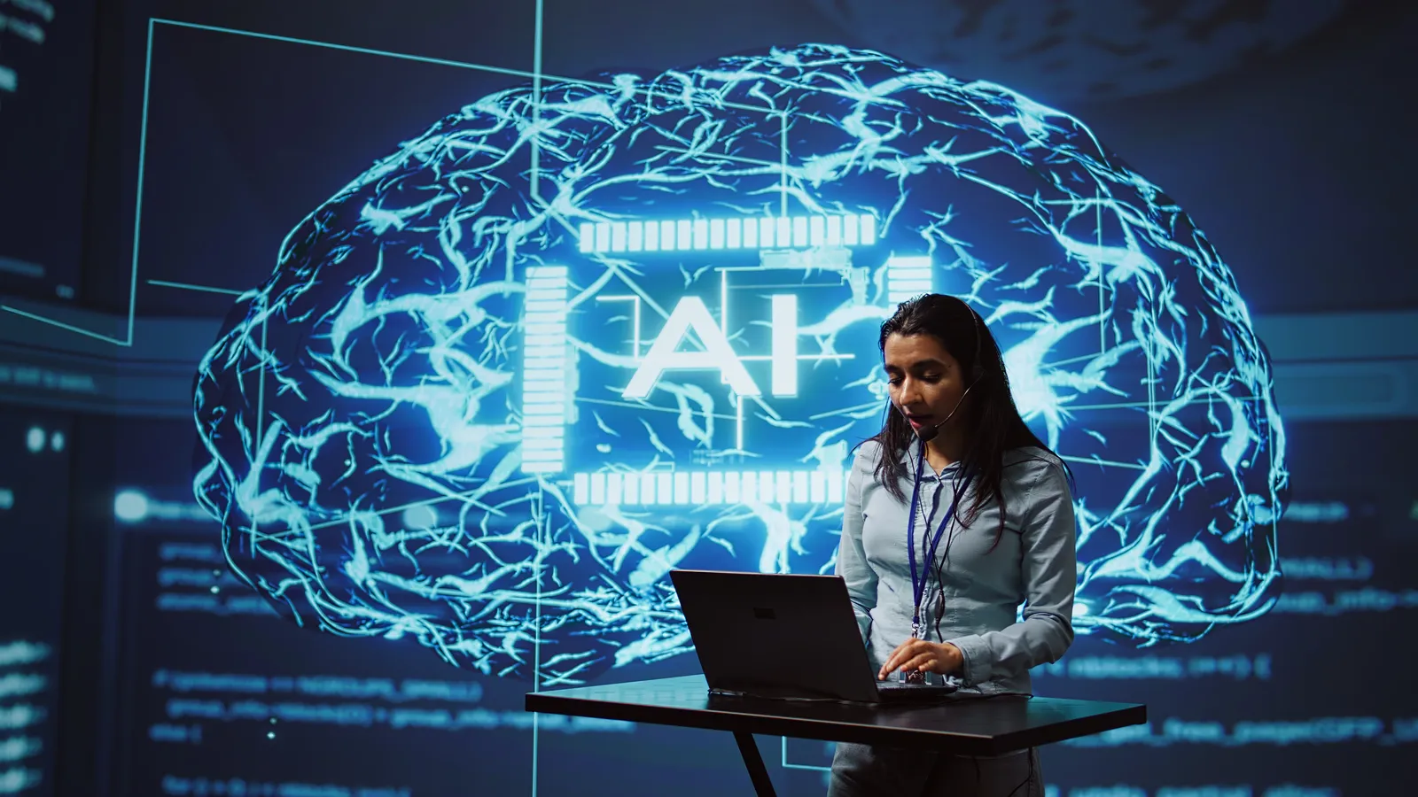 A presenter at a laptop in front of a large screen showing a glowing brain graphic labelled AI.