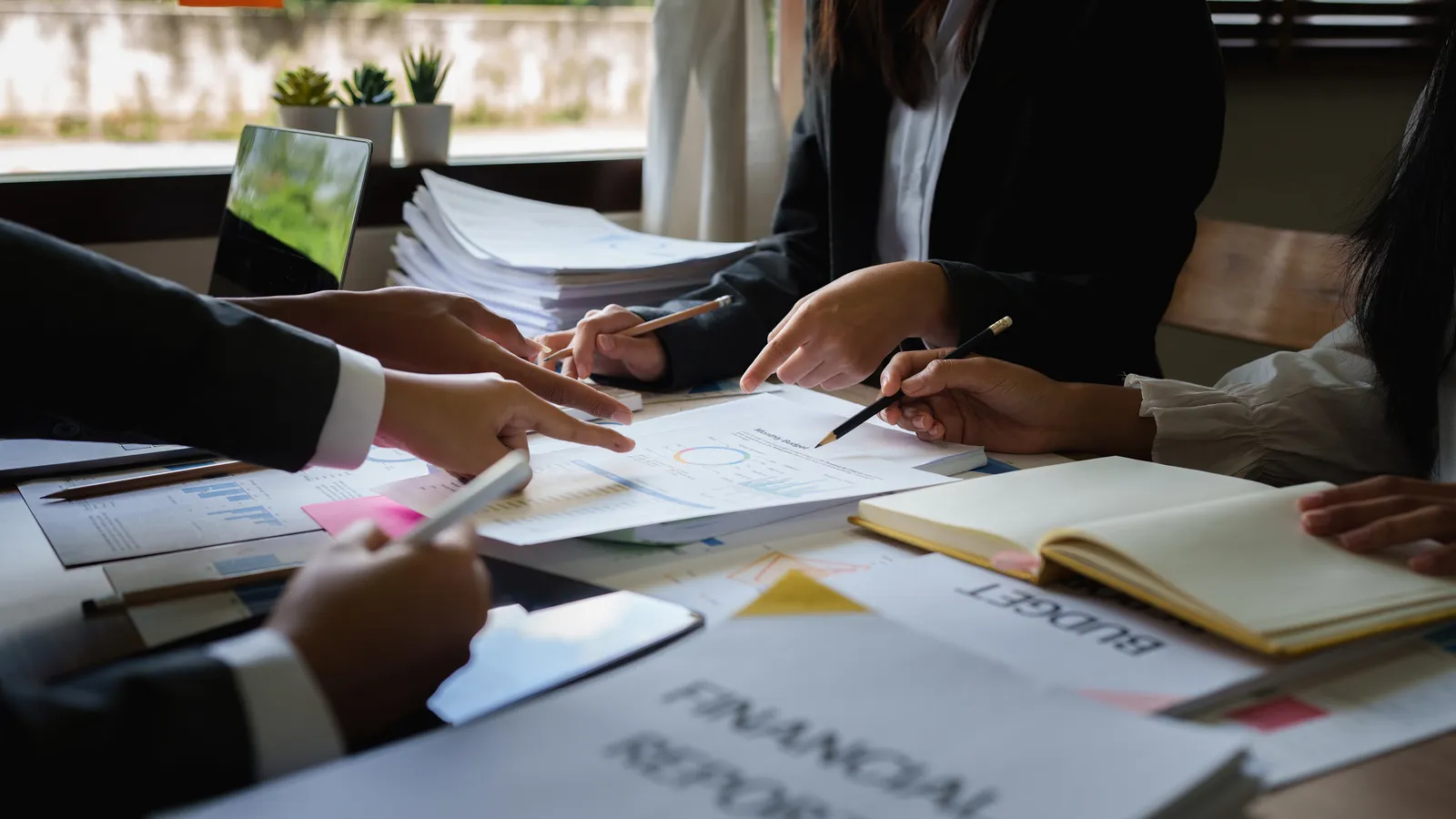Colleagues pointing at budget charts and financial reports spread across a meeting table.