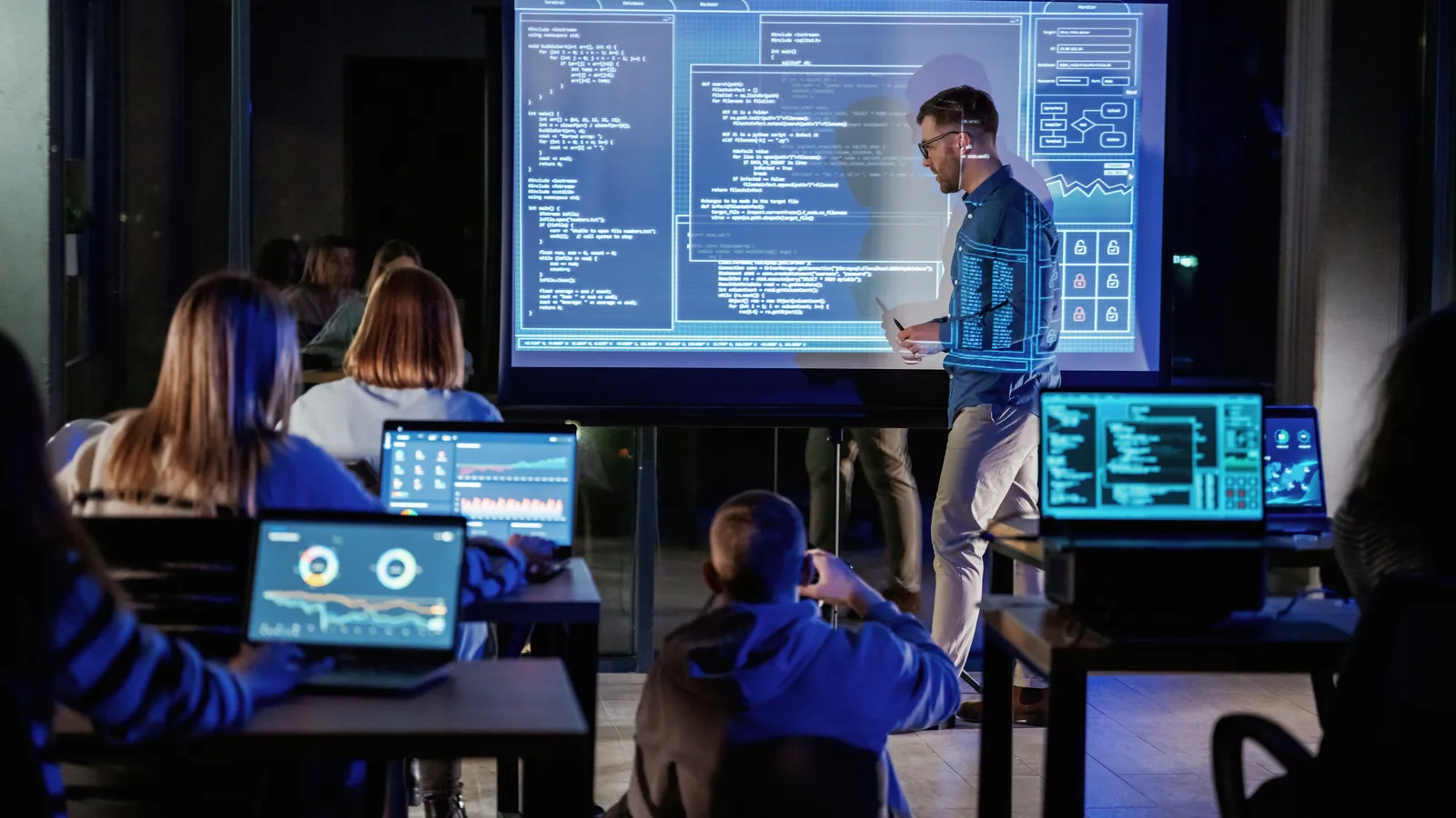 A presenter walking a room through material on a large screen while attendees follow along on laptops.
