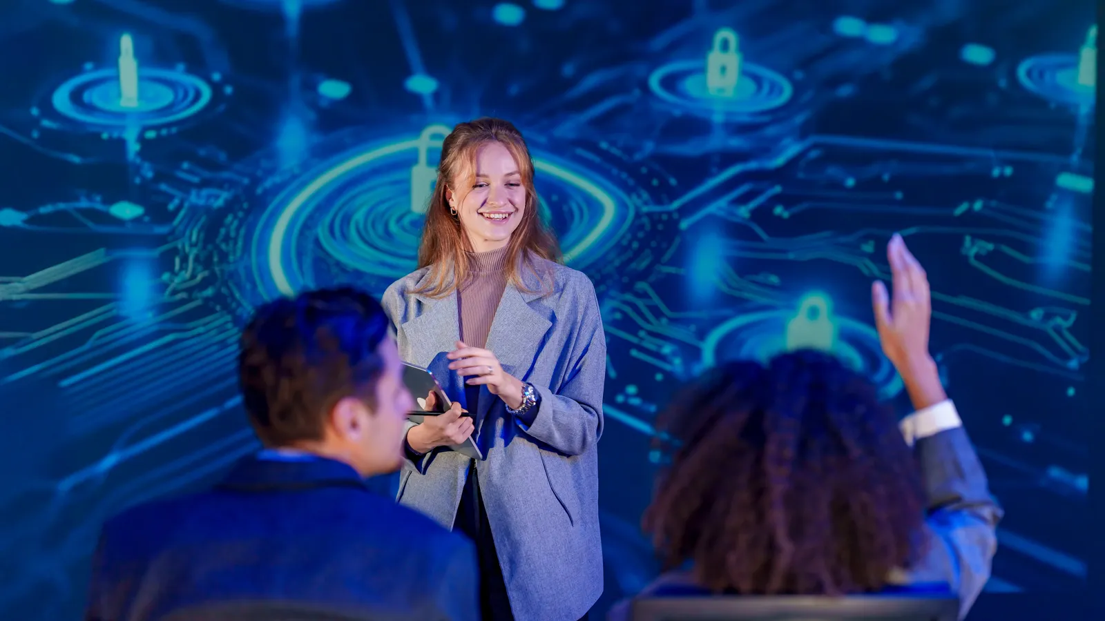 A presenter leading a cyber security training session in front of a large screen, with an attendee raising a hand.