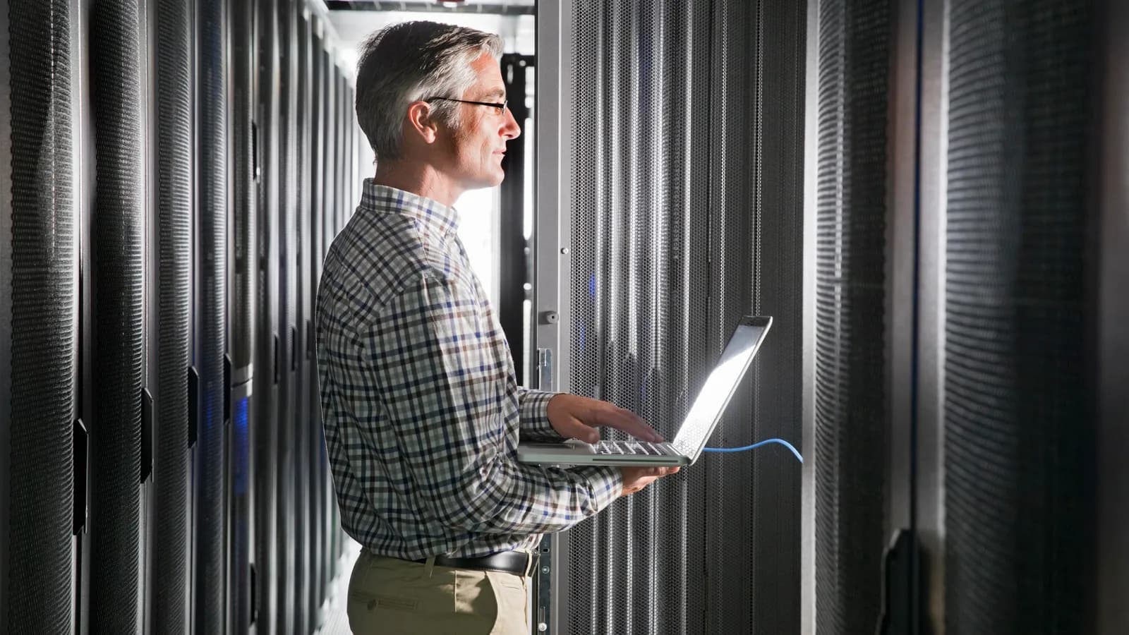 An IT professional holding a laptop connected to a server rack in a data centre aisle.